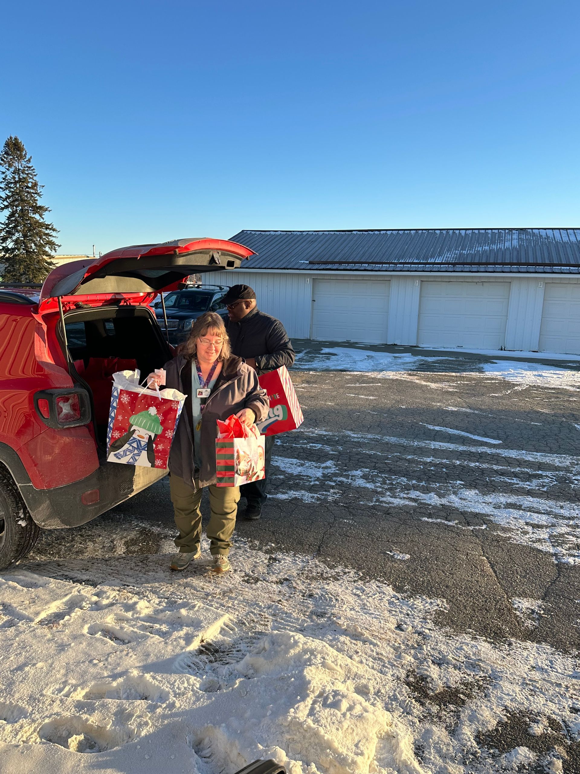 A person stands by an open car trunk in a snowy lot, holding red and white holiday-themed gift bags.