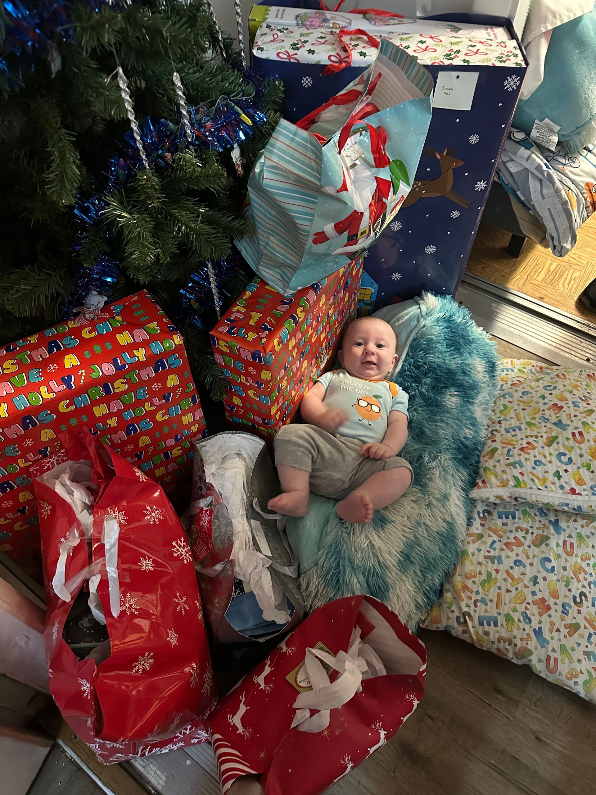 A happy infant sits amidst colorful Christmas presents under a decorated tree.