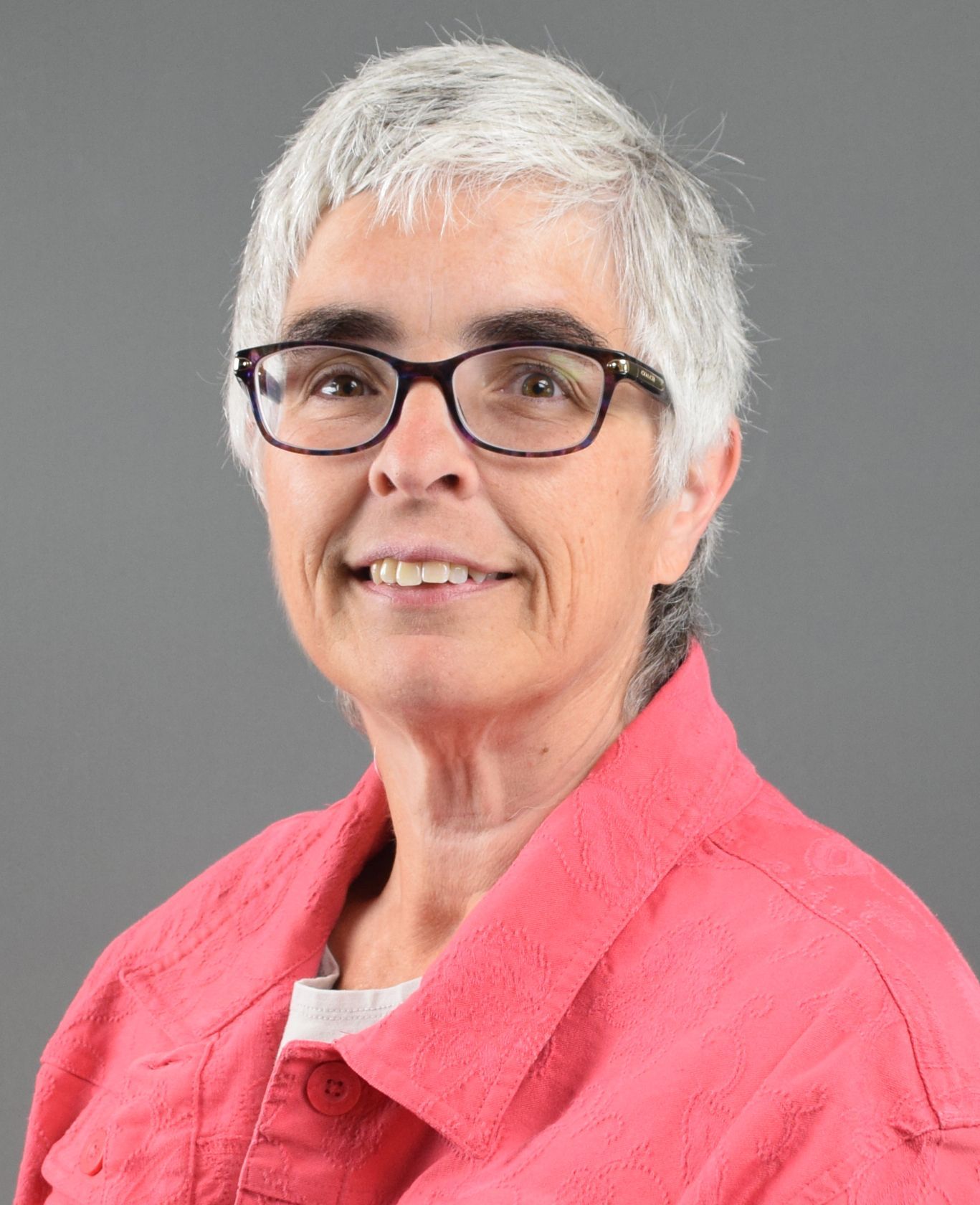A person with short, gray hair and dark-rimmed glasses smiling in front of a plain gray background, wearing a pink jacket.