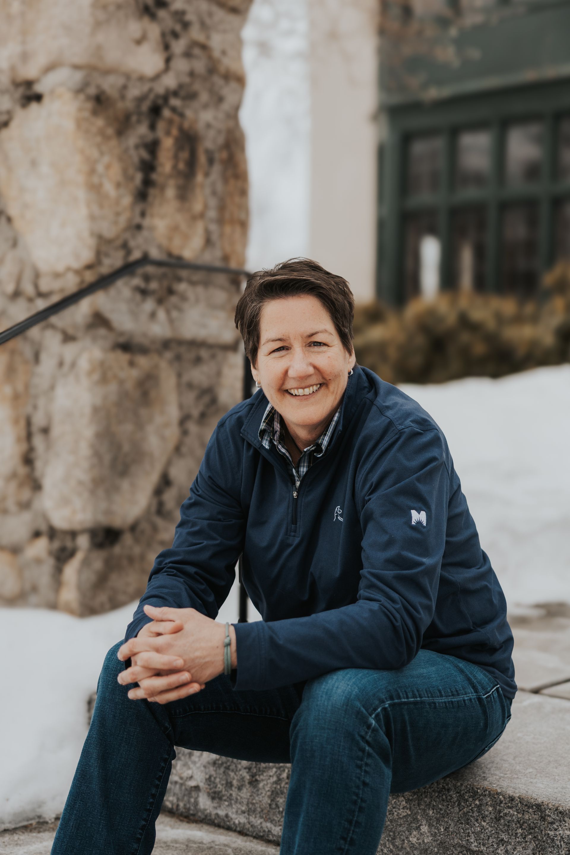 A person wearing a dark blue jacket and jeans sitting on stone steps outdoors with snow nearby, smiling at the camera.