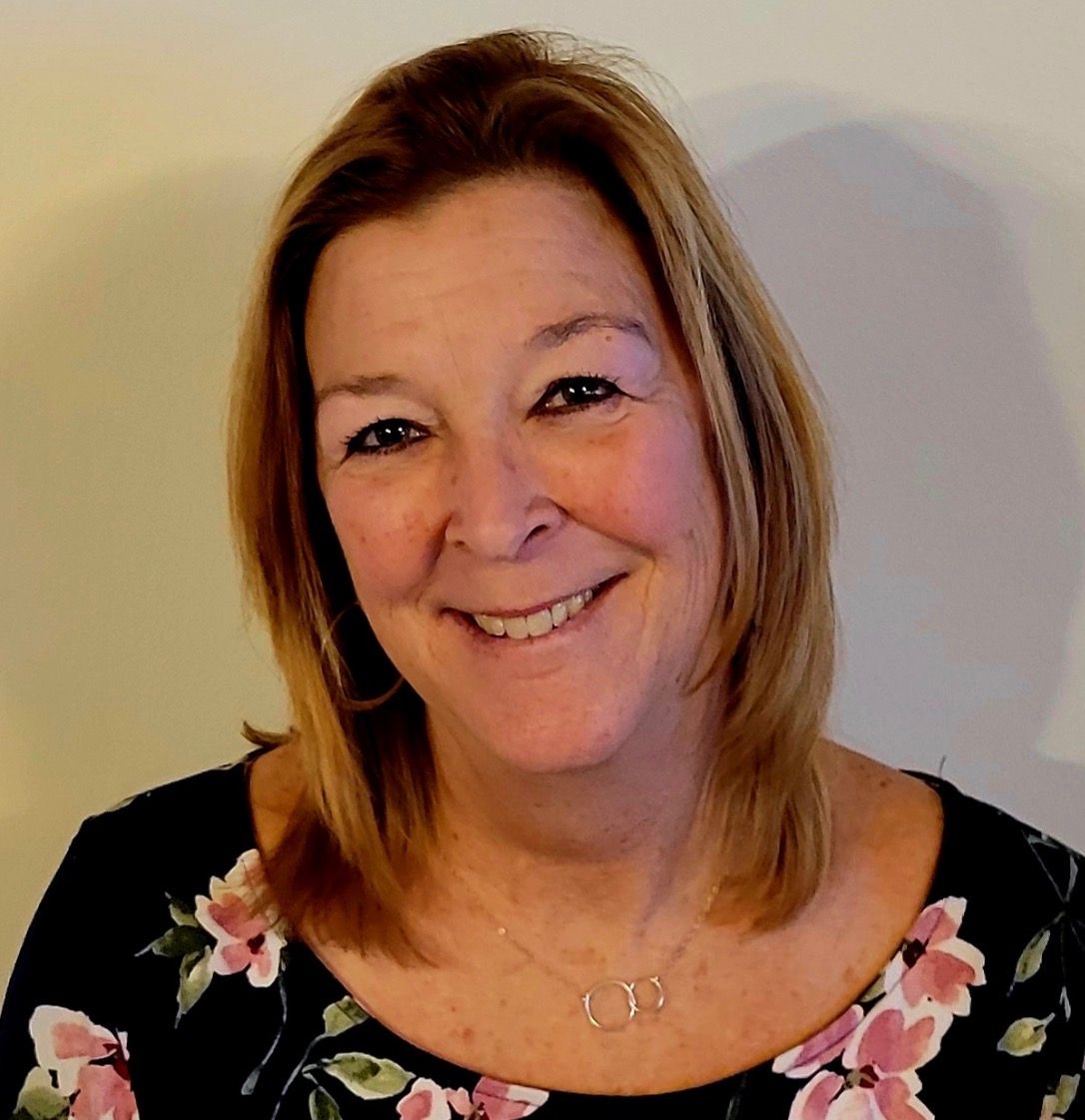 A smiling person with shoulder-length blonde hair wearing a black floral shirt and a simple necklace against a light wall.