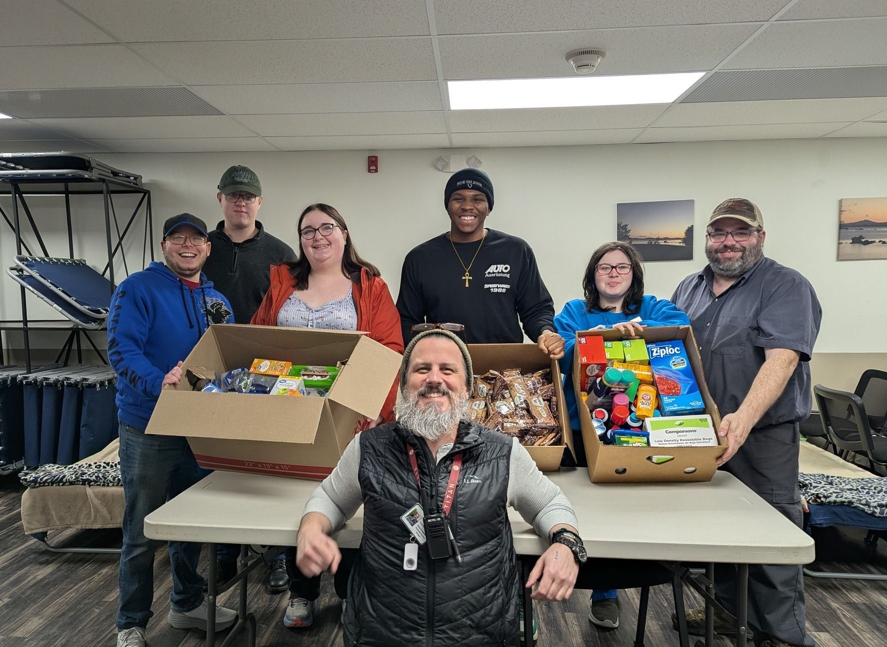 A room with four simple cots, two with blankets and pillows, set on a wood-look floor with a window covered by curtains.