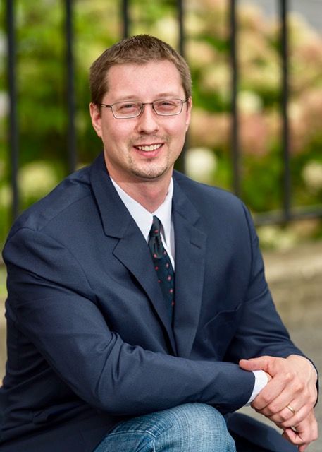 A person with glasses, wearing a blue blazer and tie, smiling while sitting outdoors near a dark metal fence.
