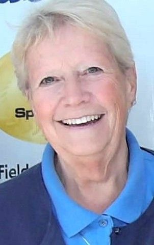 A smiling person wearing a blue collared shirt and a dark cardigan, with a blurred sign in the background.