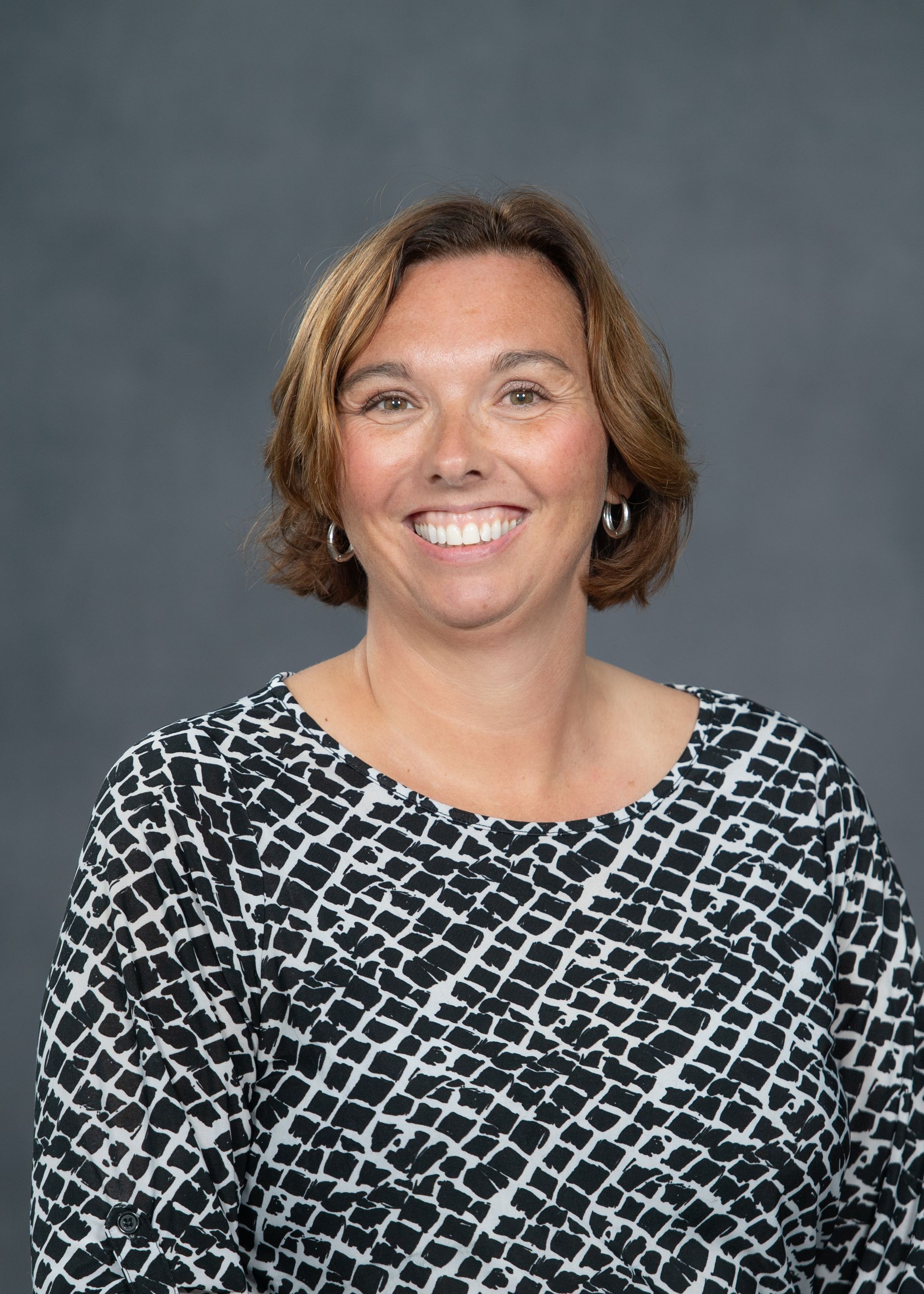 A smiling person with shoulder-length wavy brown hair wearing a black and white patterned top against a grey background.