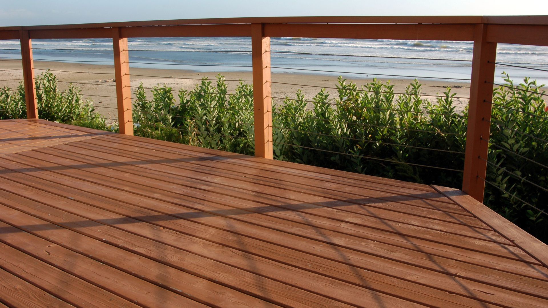 This image captures a beachfront wooden deck in Salinas, CA, offering a serene view of the ocean. The deck features warm-toned wooden planks and is bordered by a simple wooden railing, beyond which lush greenery and the sandy beach lead into the calm, expansive seascape. The deck provides a perfect spot for relaxation while embracing the coastal atmosphere, enhanced by the clear blue skies and gentle surf in the background.