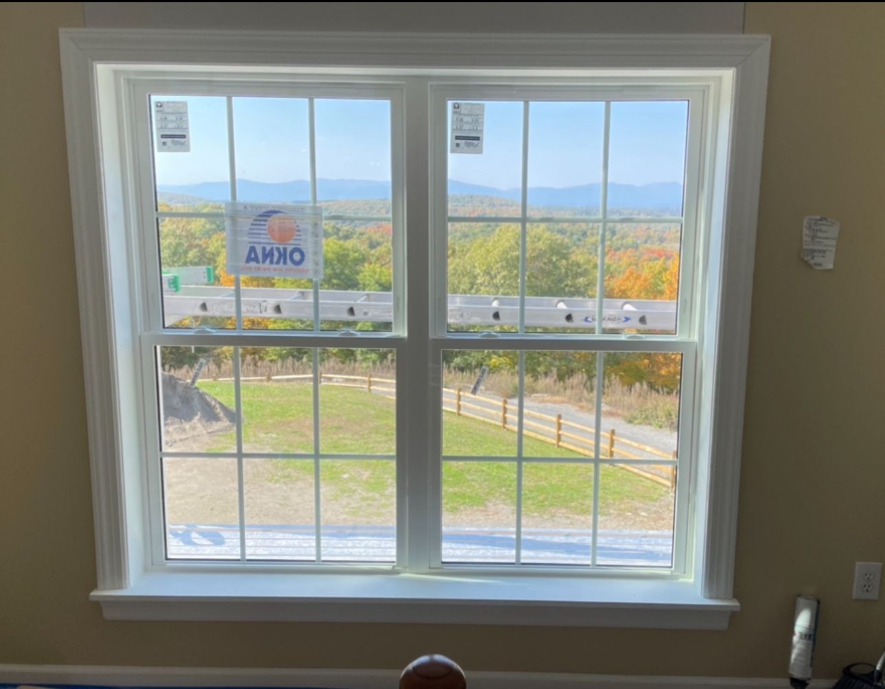 White-framed window with grid, showcasing a green lawn, wooden fence, and distant mountains under a blue sky.