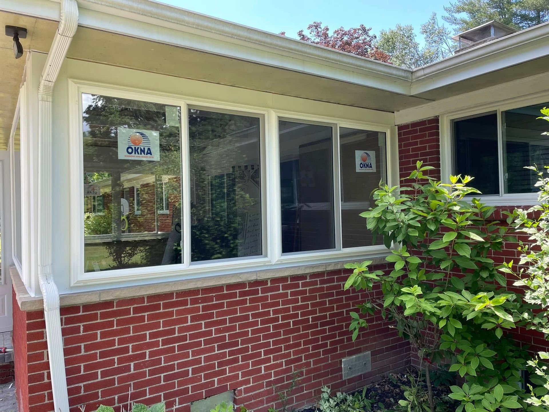Brick house exterior with white-framed windows, surrounded by greenery under a bright sky.