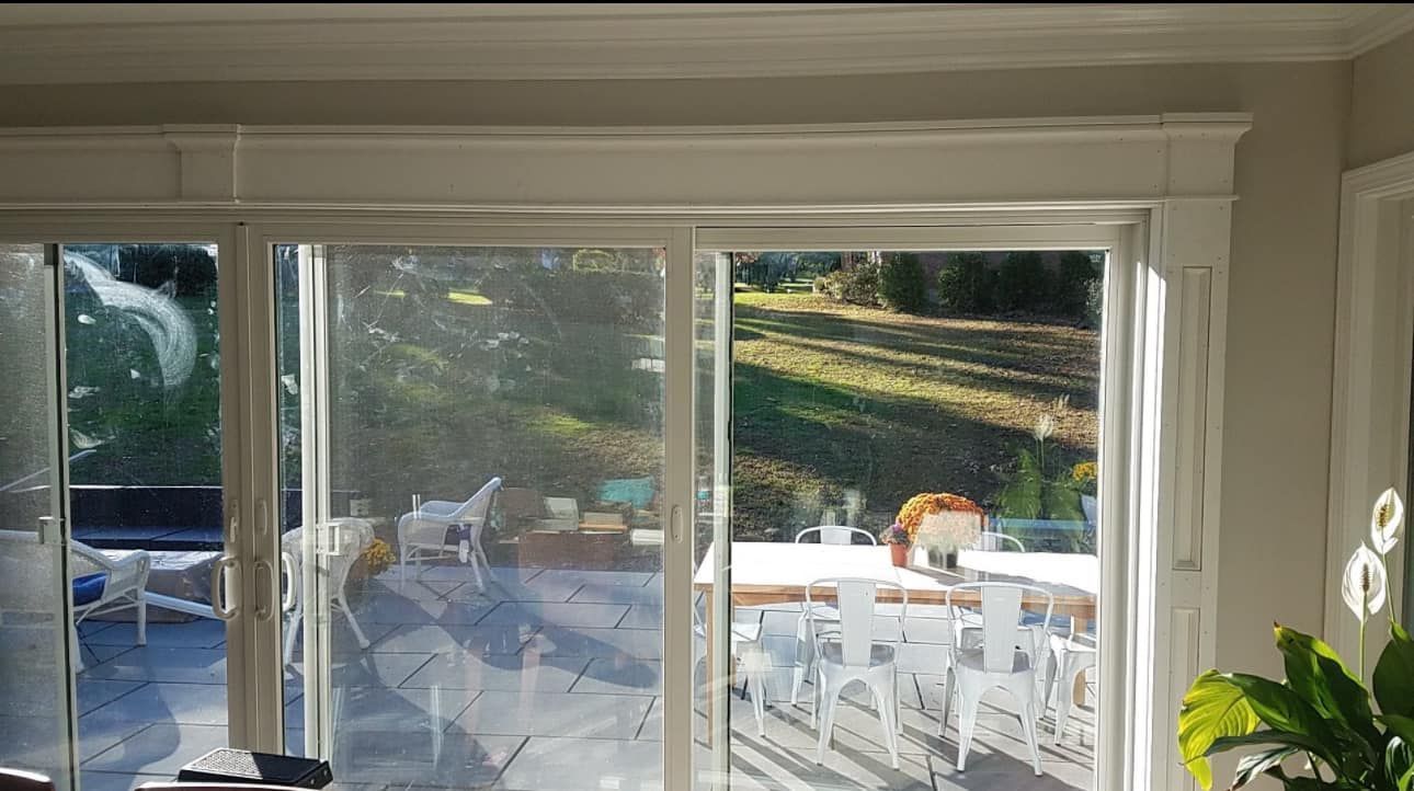 Sliding glass doors open onto a patio with white chairs and a backyard. Sunlight streams in.