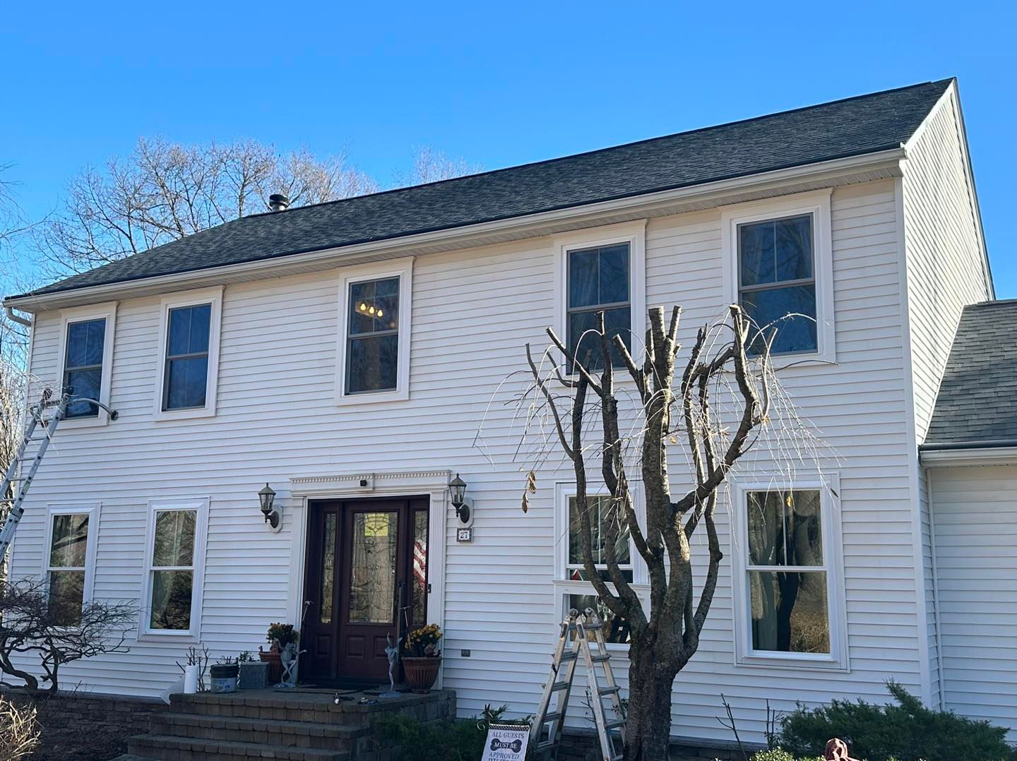 White two-story house with dark roof and multiple windows under a blue sky. A ladder rests against the building.