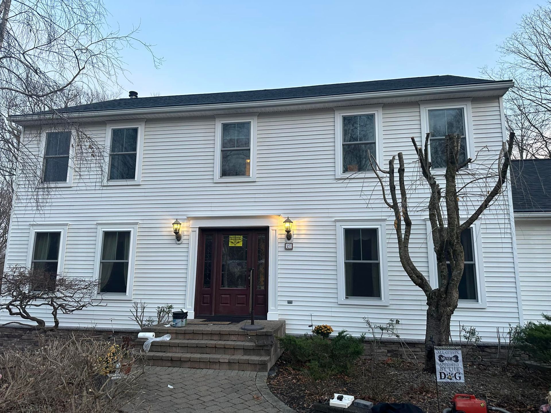 Two-story white house with dark door and windows. Bare tree and shrubs in front. Steps lead to entrance.