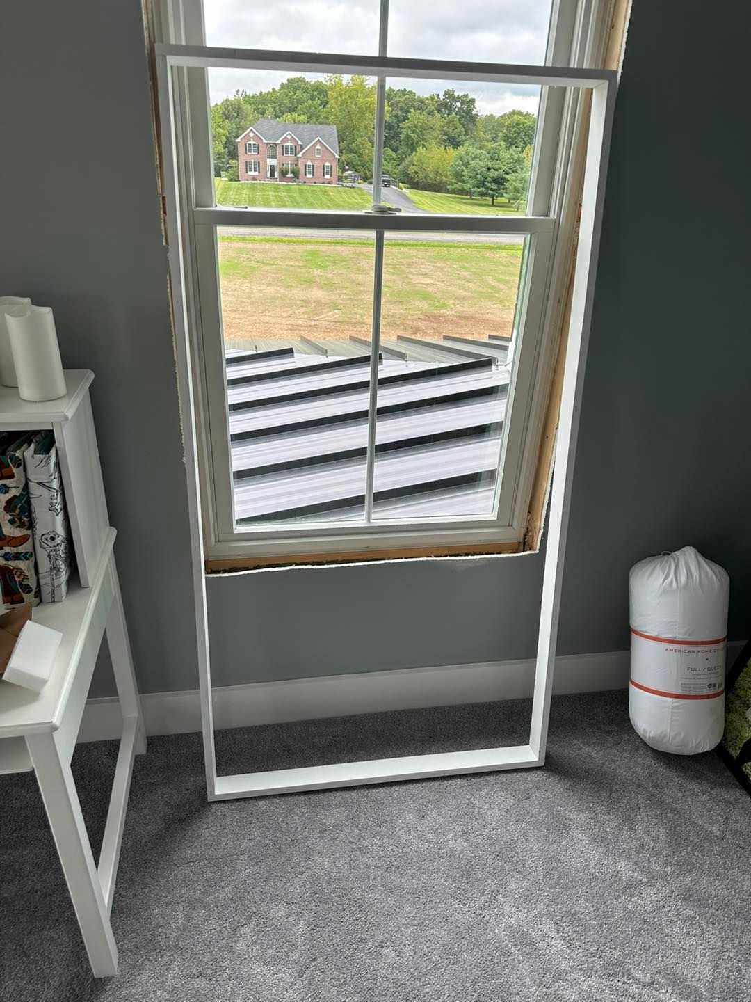 White window frame leaning against a wall next to an open window overlooking a house and trees.