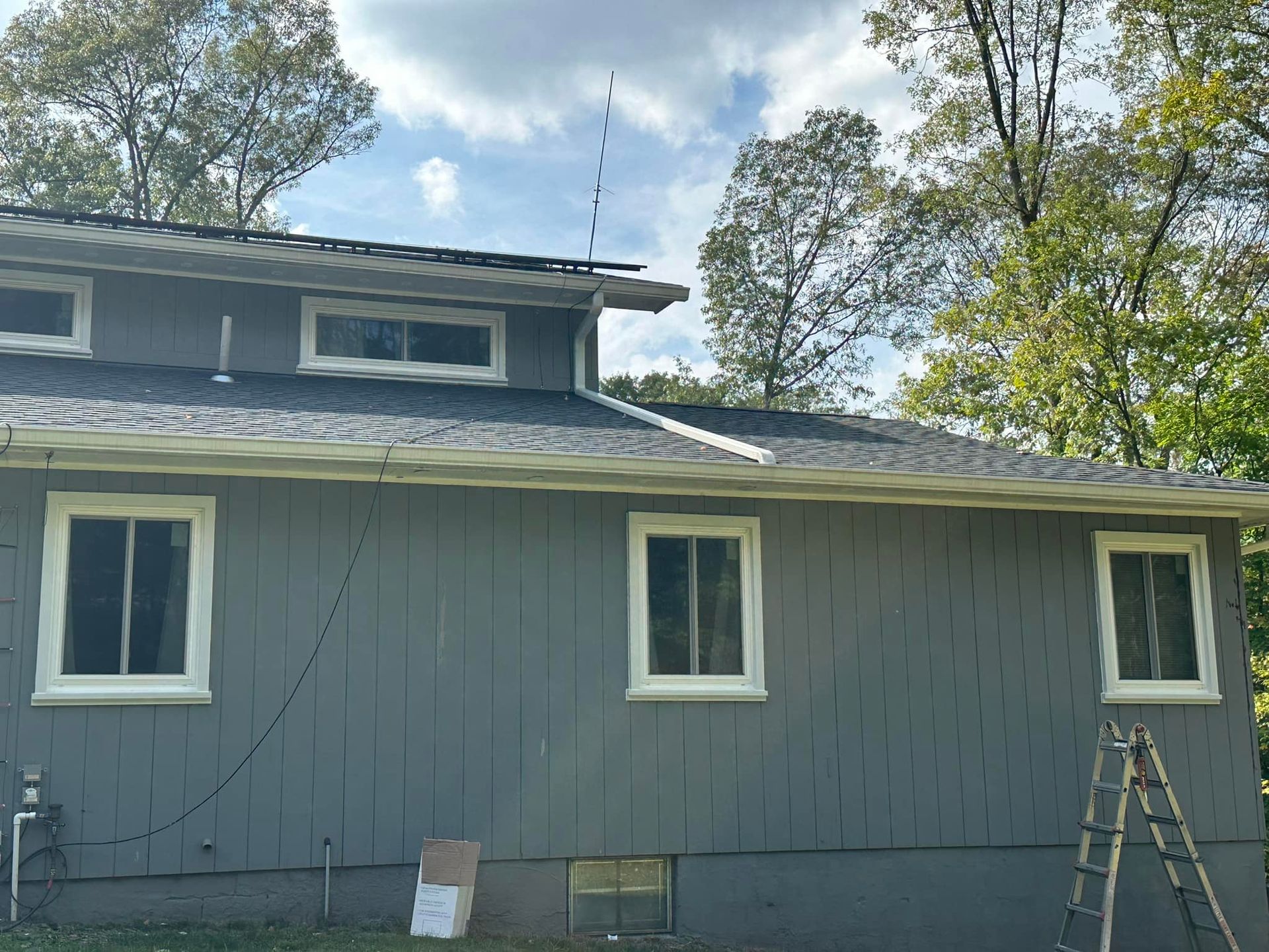 Gray house exterior with white-framed windows, dark roof, trees, and ladder.
