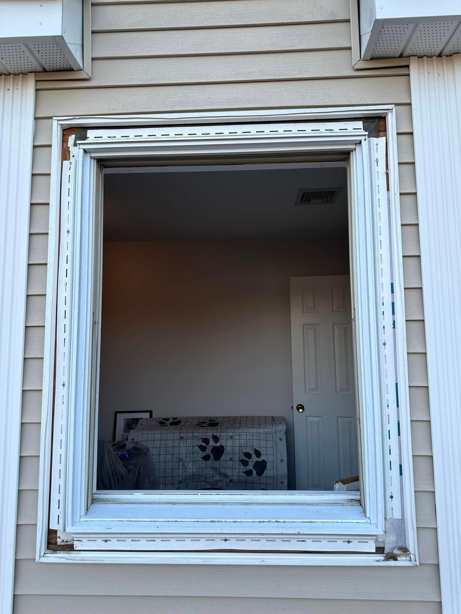 Exterior view of a window frame, missing glass. Shows interior: a room with a closed door, a pet carrier, and a painting.