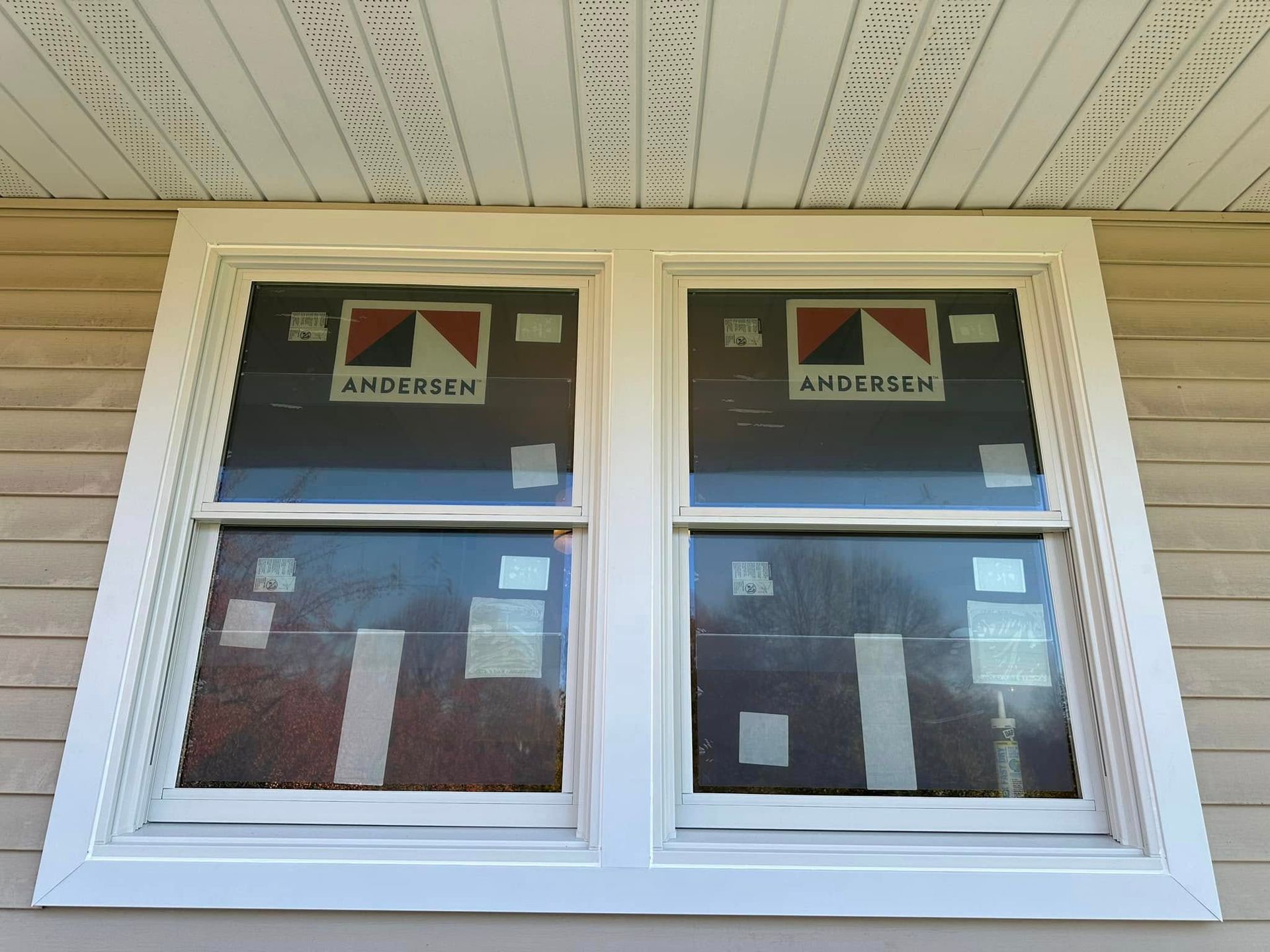 Two white framed windows with new Andersen window coverings, installed in a beige siding exterior.