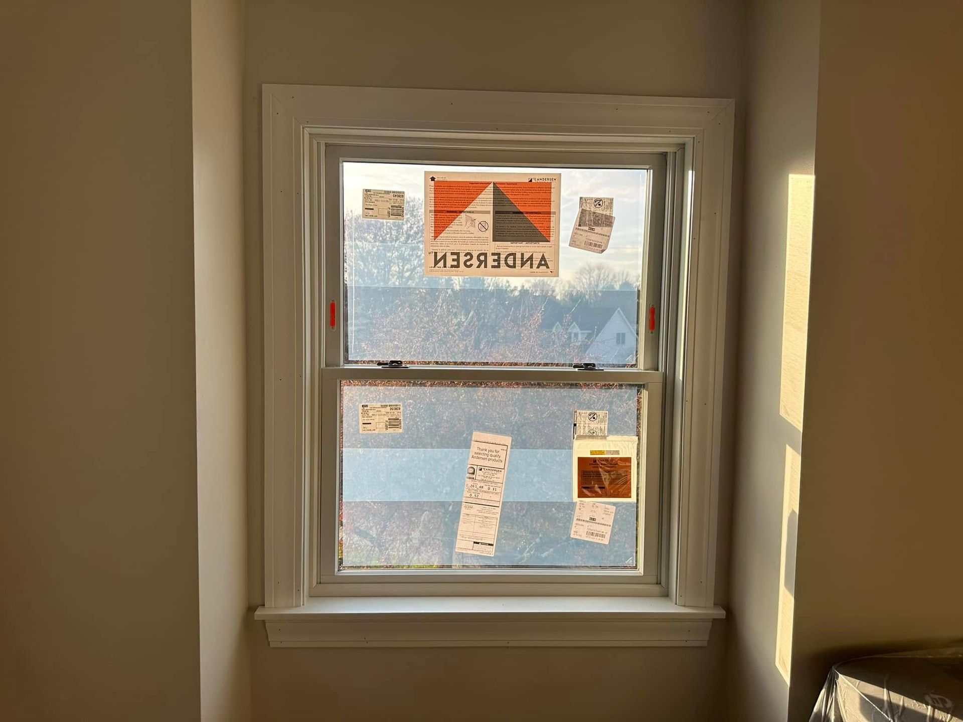 Window with new Andersen branding stickers, reflecting a bright, hazy outdoor scene.