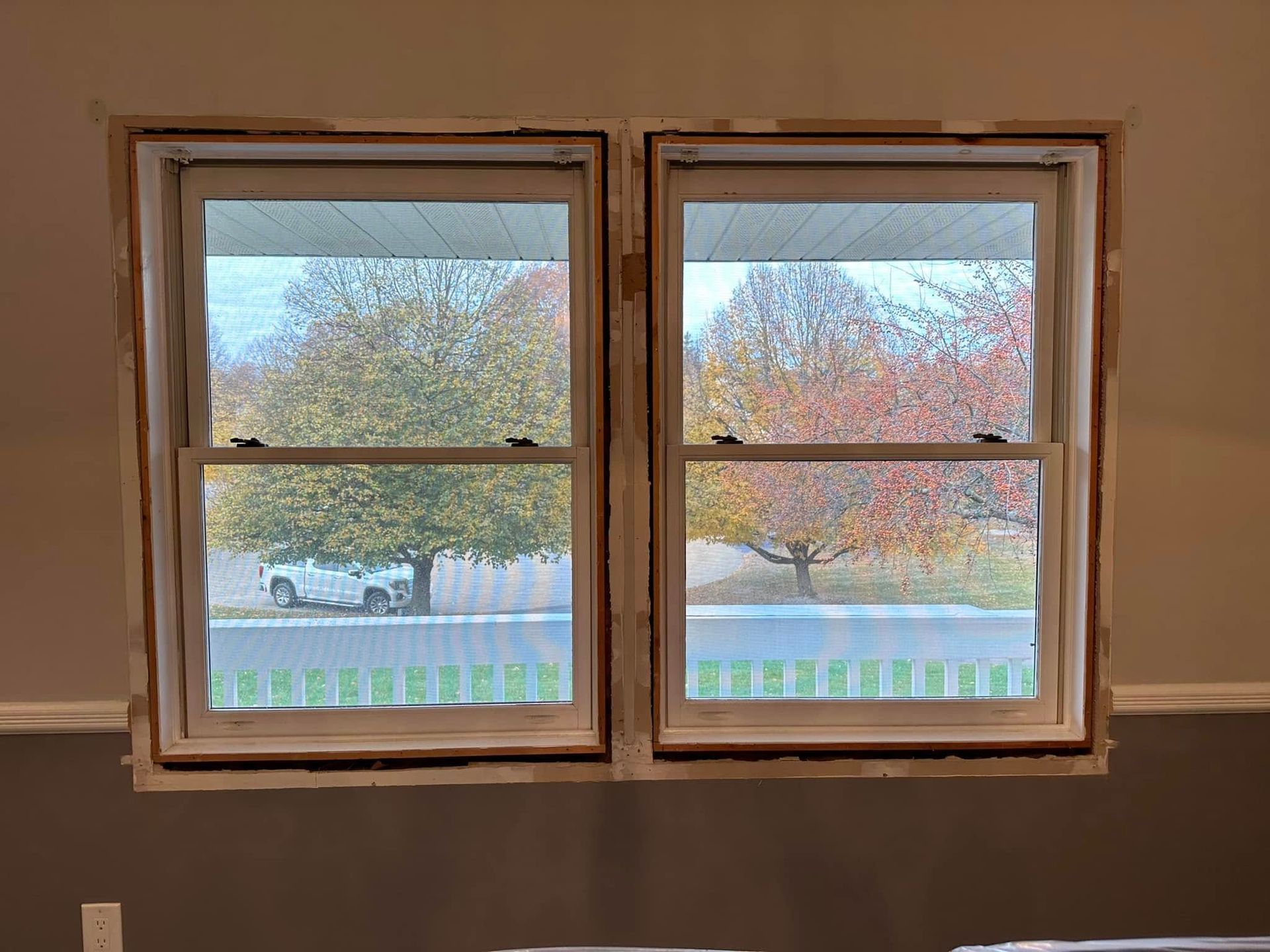 Two double-hung windows showing a view of colorful autumn trees and a street from a room with unfinished wall trim.