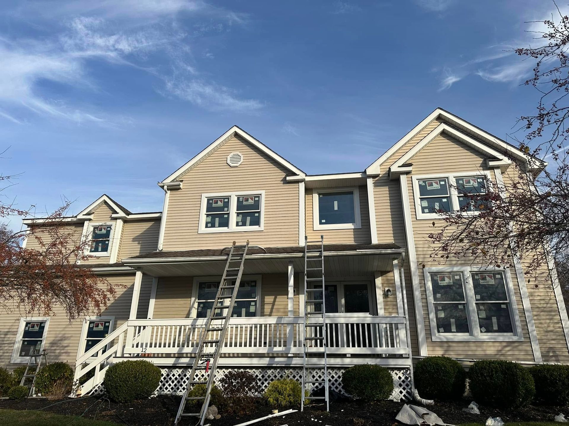 Two-story house with beige siding, white trim, and ladders. Windows are visible, under a blue sky.