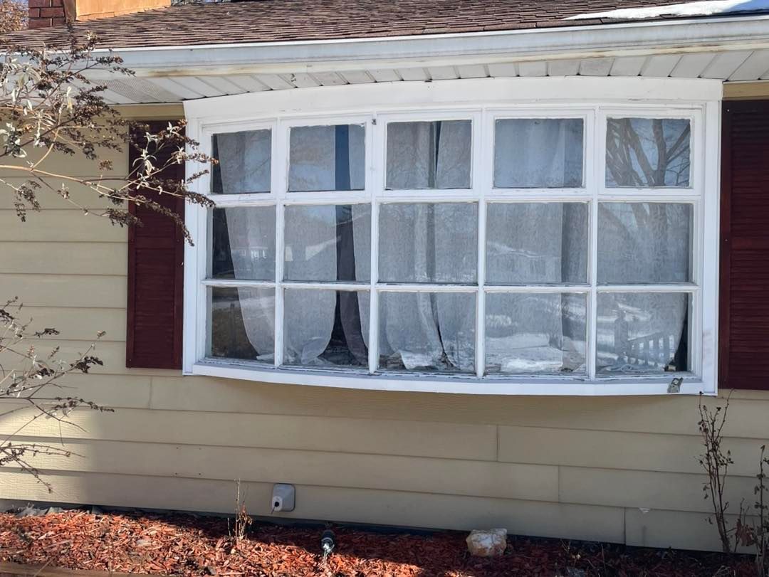 Bay window of a house with white trim, tan siding, and red shutters; curtains are visible behind the glass.