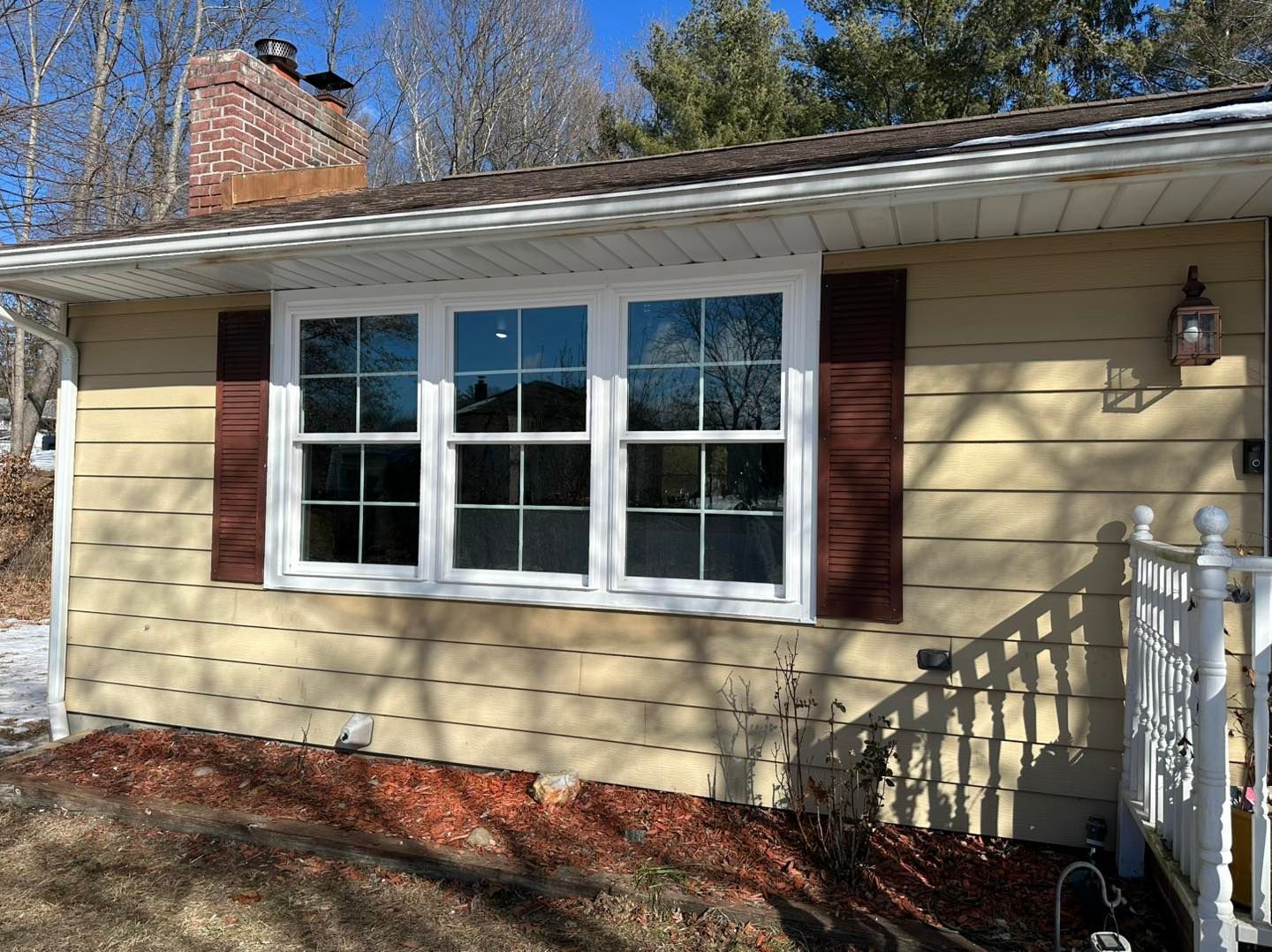 Tan house exterior with three white-framed windows, brown shutters, and a brick chimney.