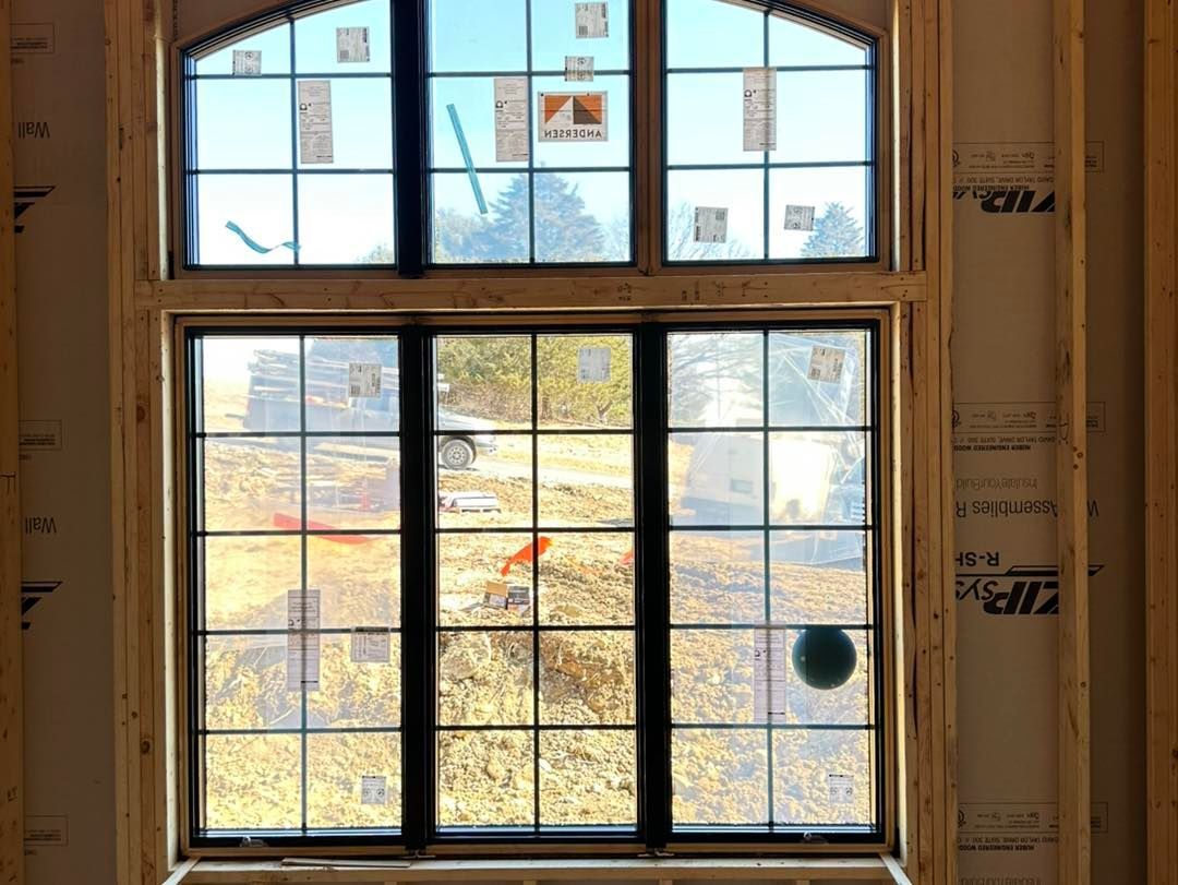 Large window with black grid panes, framed by unfinished wooden walls, looking out onto a sunny, grassy field.