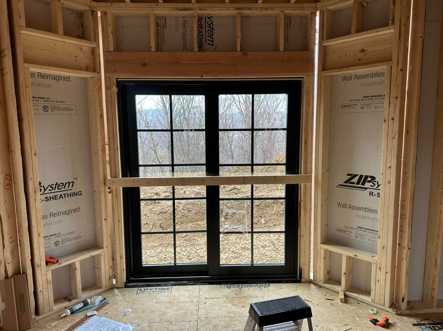 Interior framing around a black-framed window. Wooden structure with a view of a wooded area outside.