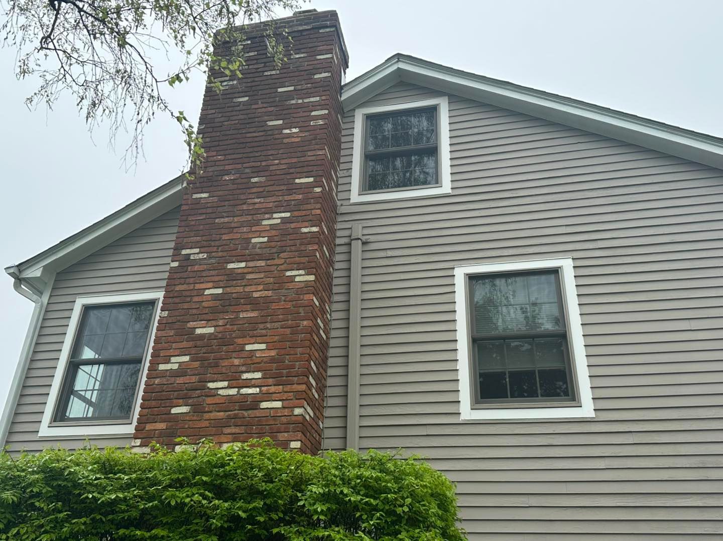 Brick chimney next to a gray-sided house with three windows. A green bush is below.