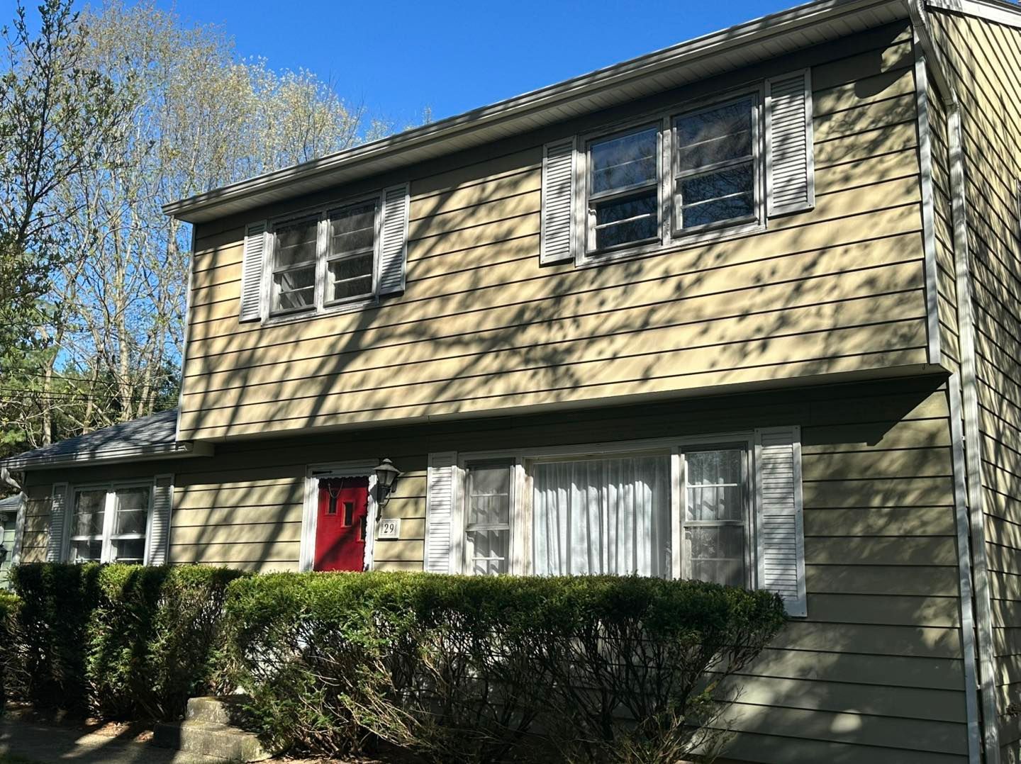 Two-story house with tan siding, white shutters, and a red front door. A green hedge is in front.