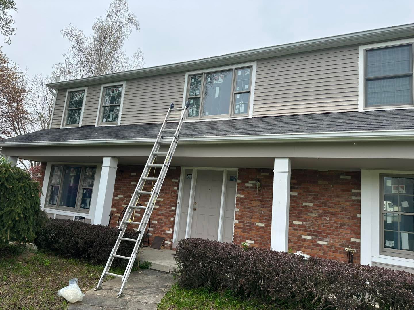 Two-story house with a ladder propped against the roof. Gray siding, brick facade, windows, and bushes. Overcast sky.