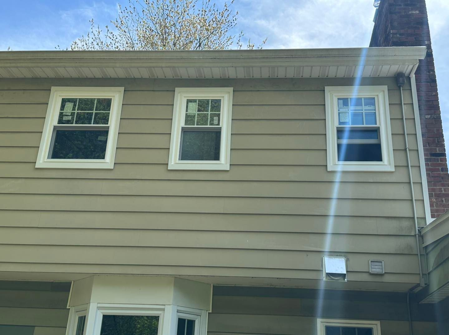 Three white-framed windows on the beige siding of a house with a brick chimney and blue sky.