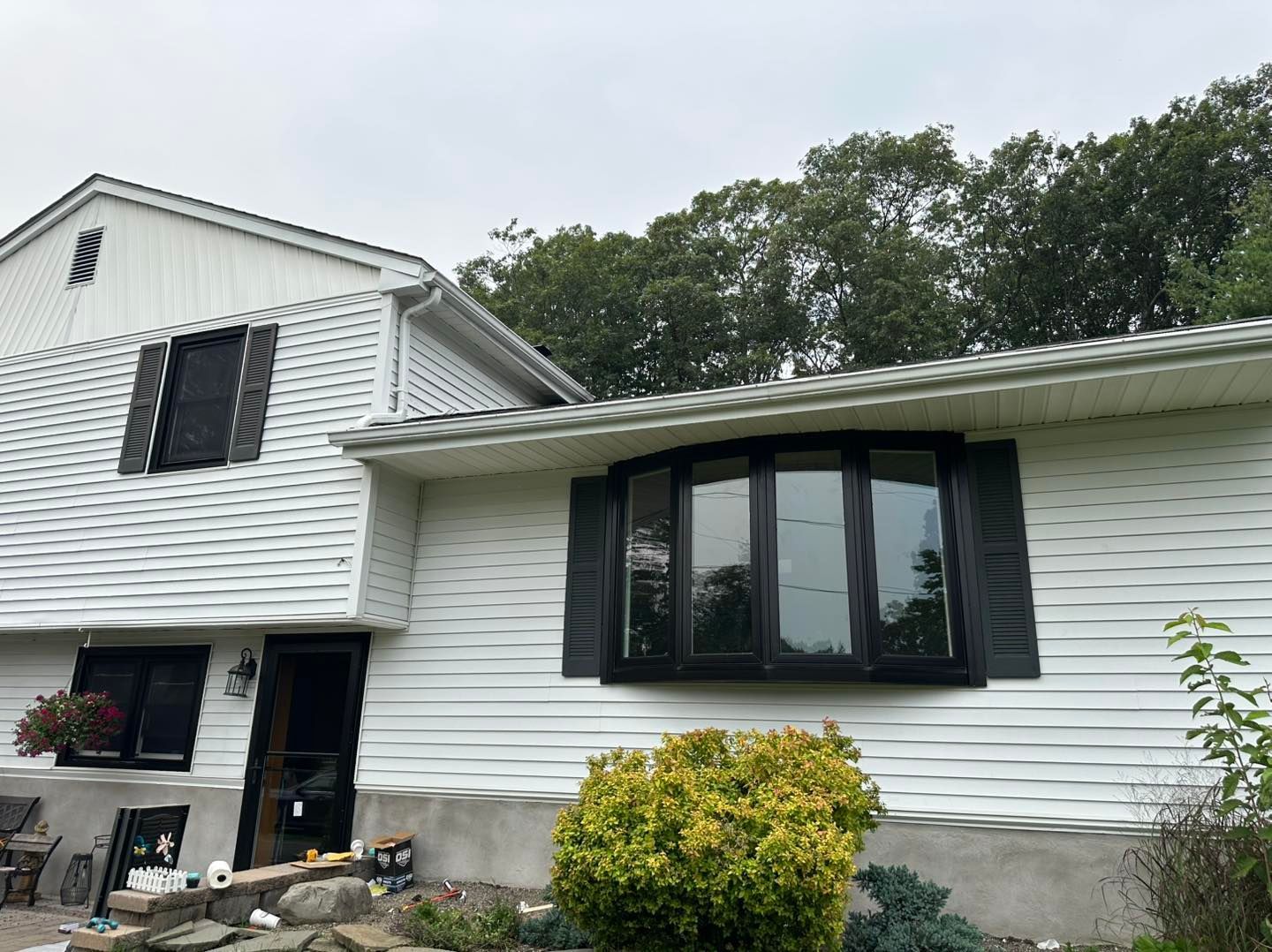 White house with black shutters and a bay window, set against a cloudy sky and trees.