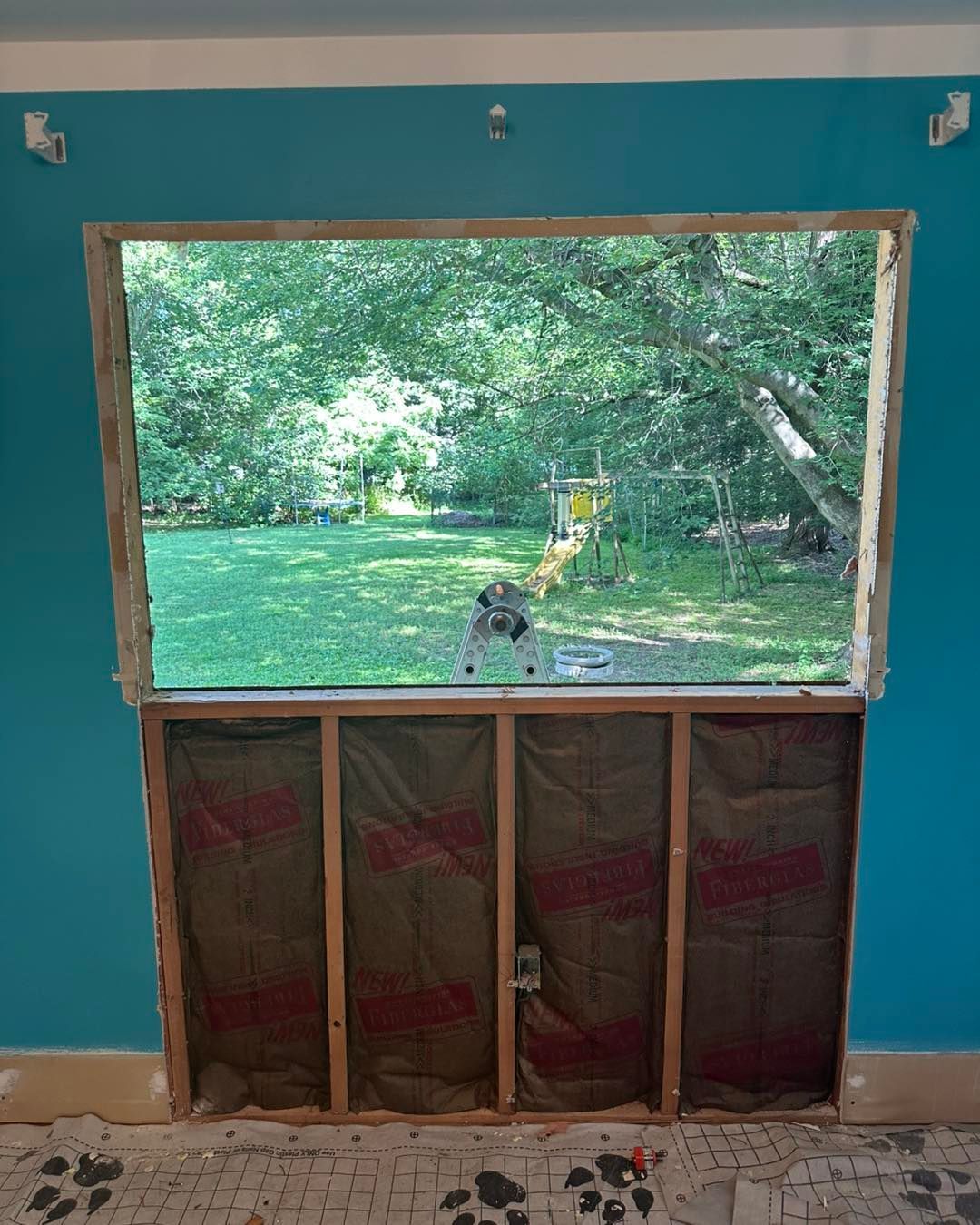 Interior view of a window frame being rebuilt. Framing, insulation, and teal wall are visible. The outside features greenery.
