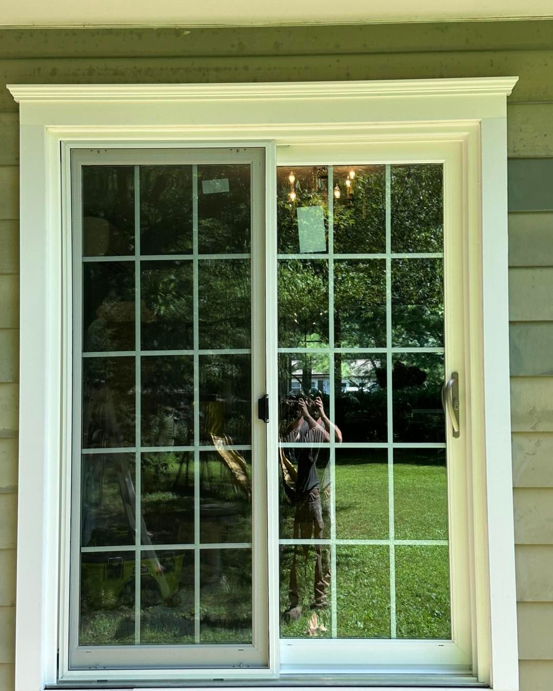Sliding glass door with white grid patterns, reflecting a green yard and trees. A person is visible in the reflection.