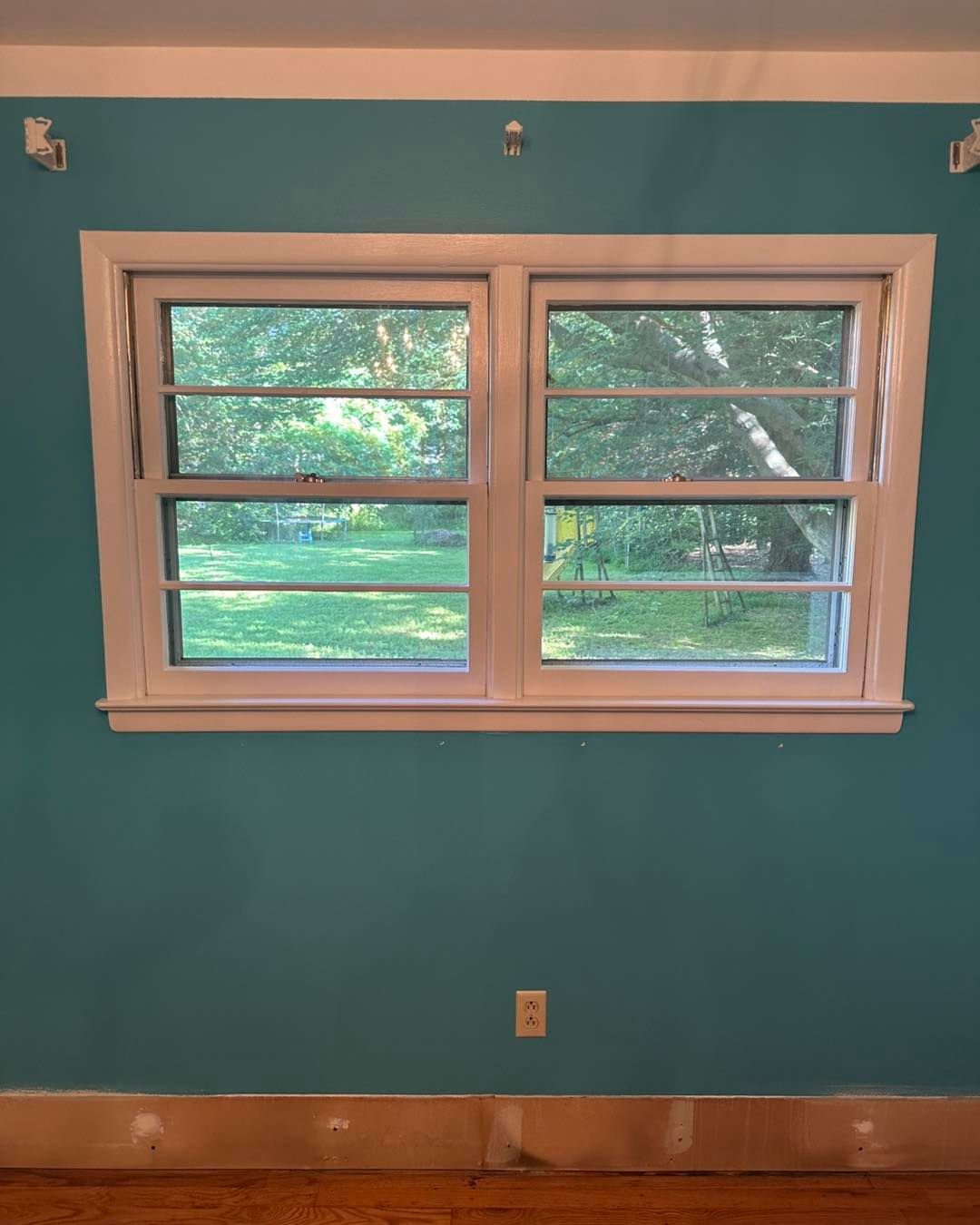 White-framed double-hung window on a teal wall, looking out onto a green yard with trees.