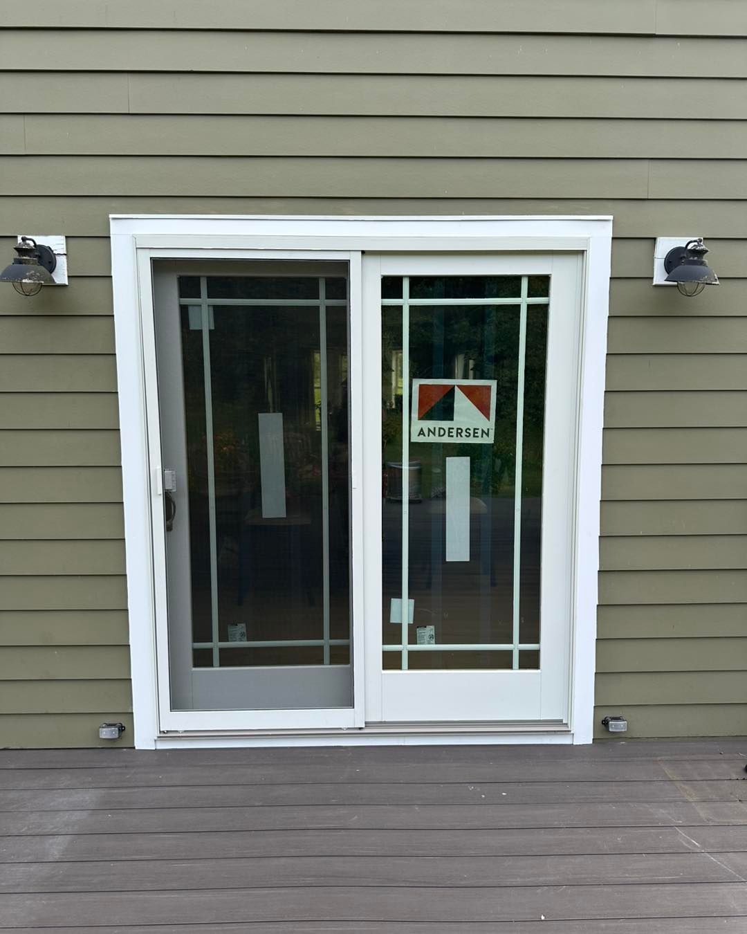 Sliding glass door on a wooden deck, flanked by lights, with green siding background.