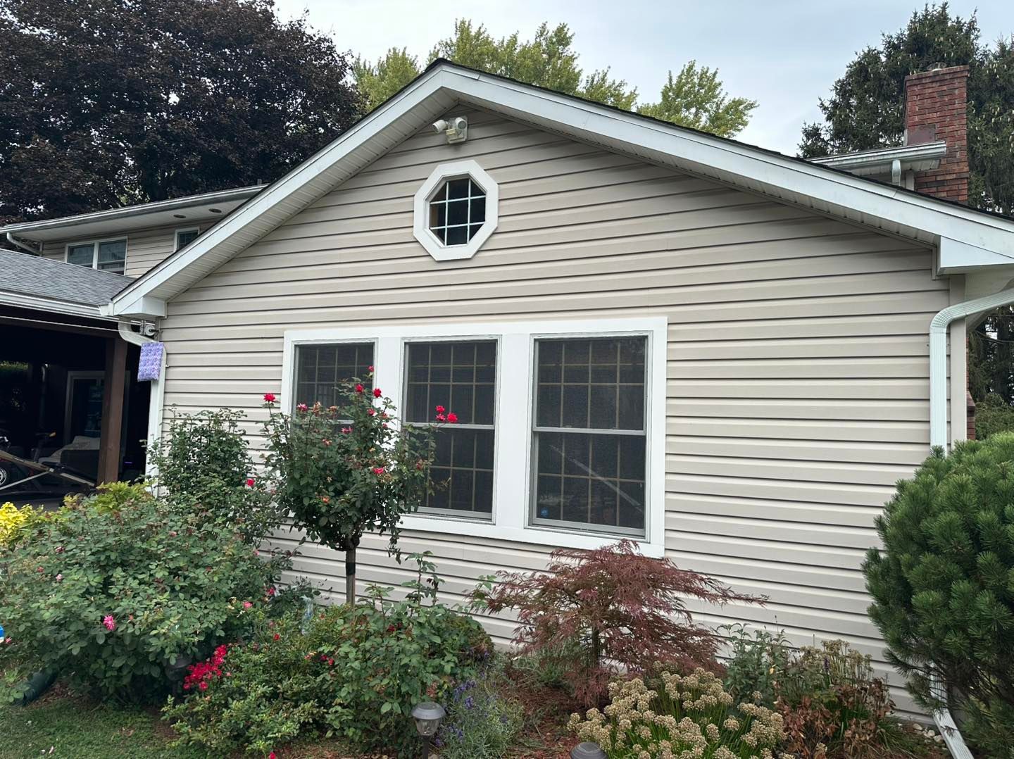 Beige house with three windows and an octagonal window above, surrounded by greenery.
