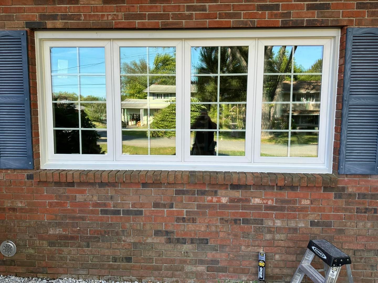 Brick house with white-framed window reflecting trees and a person. Blue shutters flank the window.