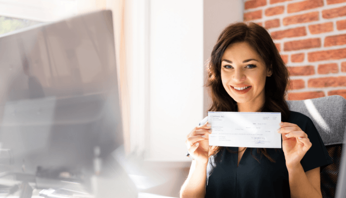 Young professional woman smiling at her desk, holding her paycheck.