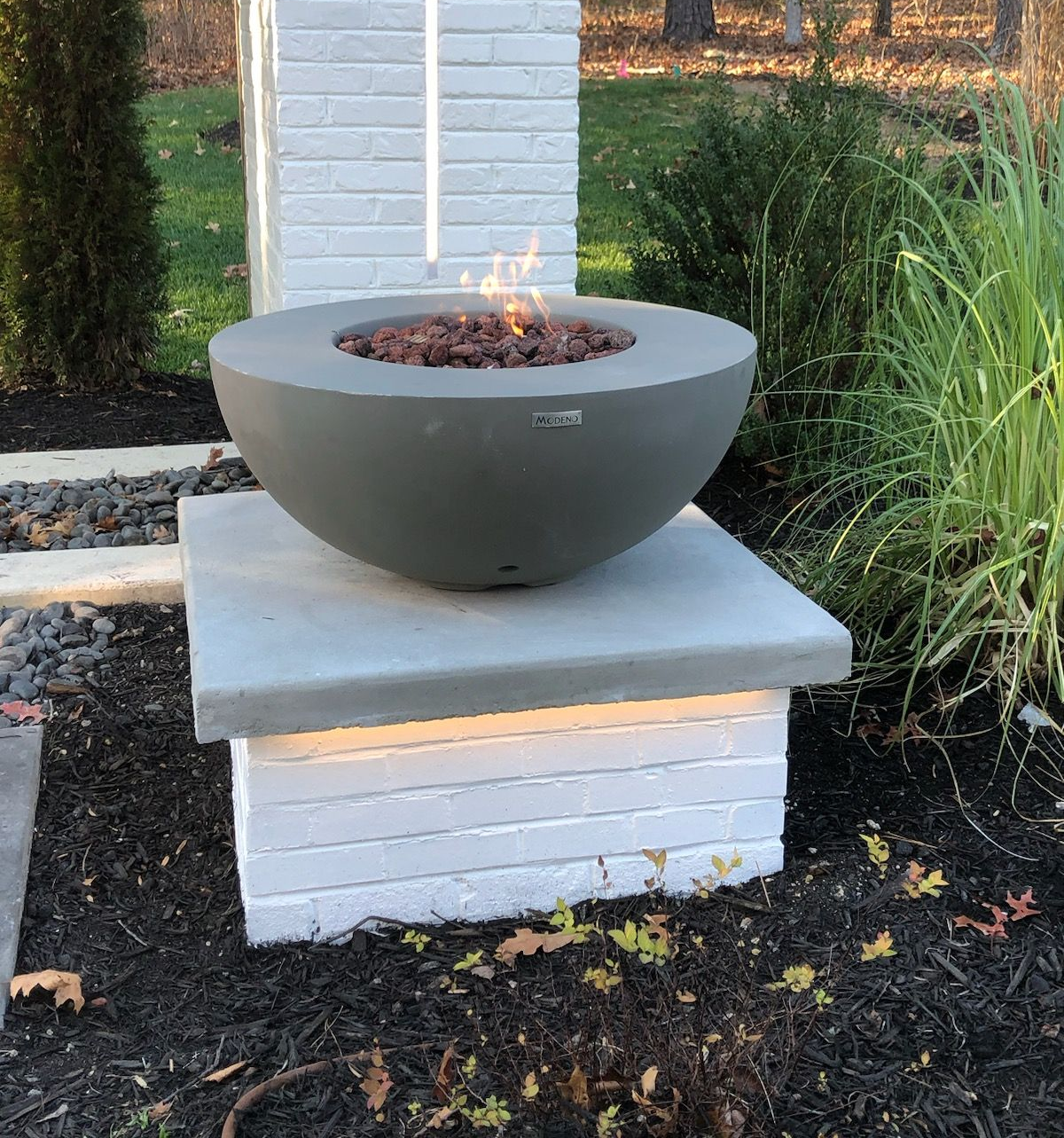 Fire pit in a gray bowl shape, sits on a white brick pillar with glowing lights, surrounded by landscaping.