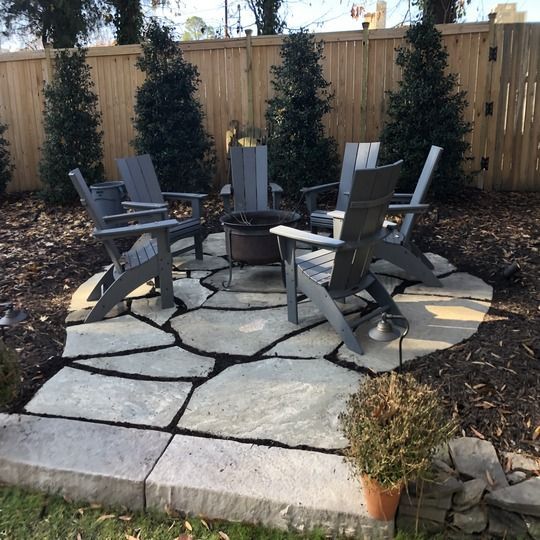 Outdoor seating area: gray chairs around a fire pit on stone pavers, surrounded by mulch and greenery.