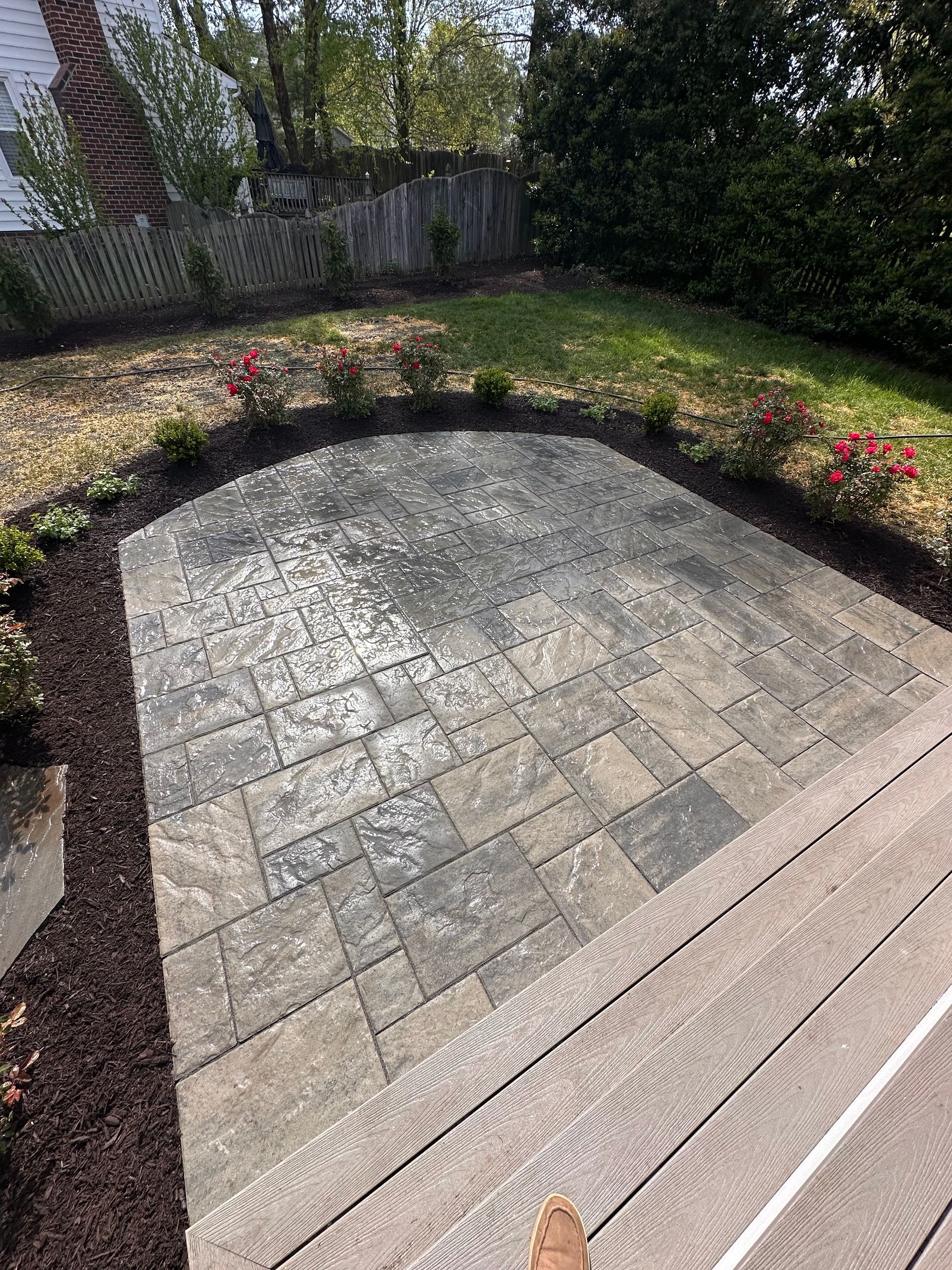 Paver patio in backyard with surrounding mulch, grass, and foliage.