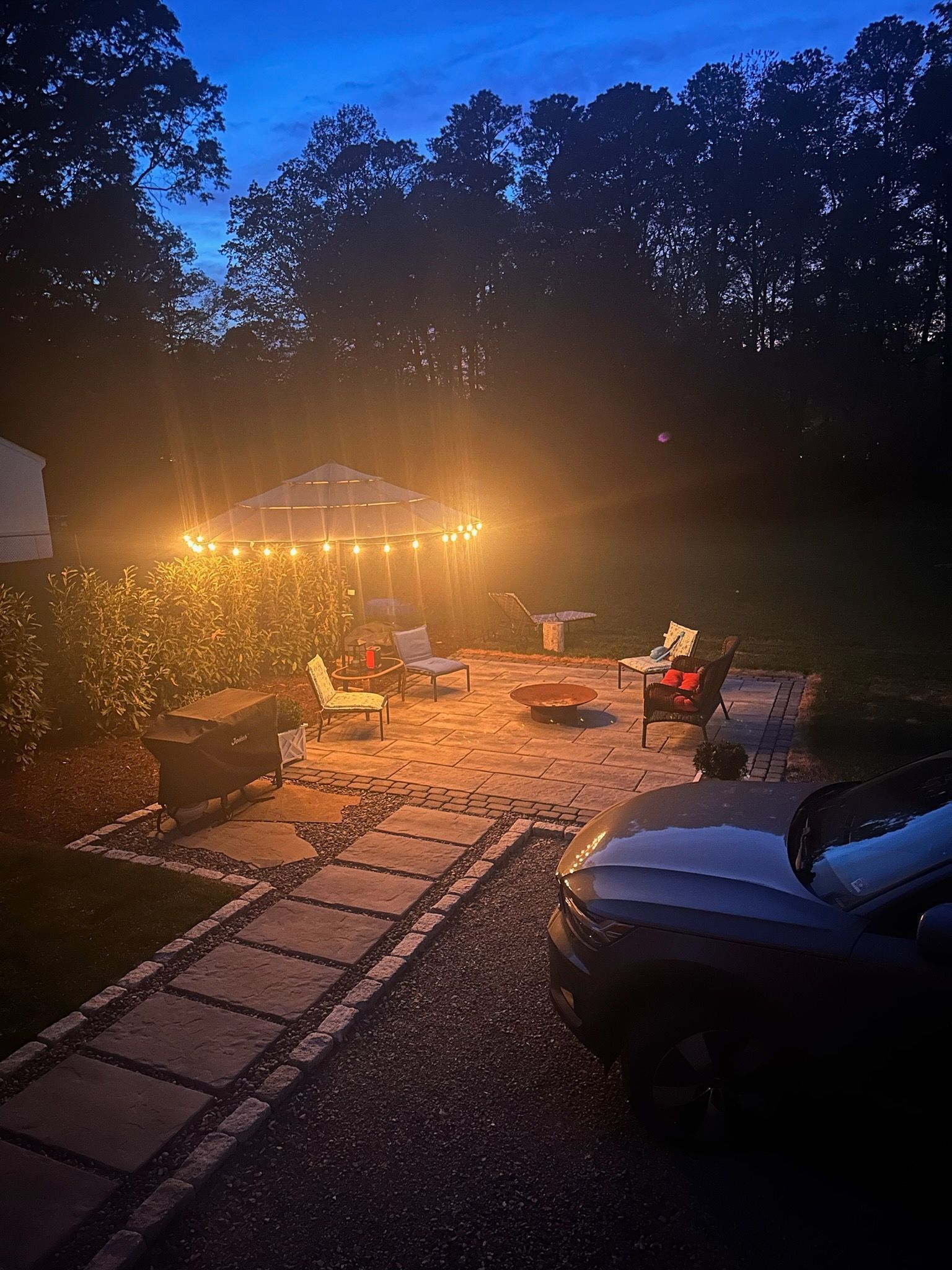 Patio at dusk lit by string lights; outdoor furniture, fire pit, and a dark car.