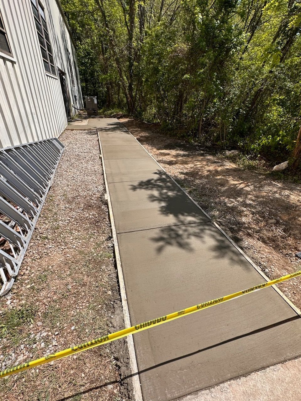 Freshly poured concrete sidewalk next to a building, yellow caution tape in foreground, trees in the background.