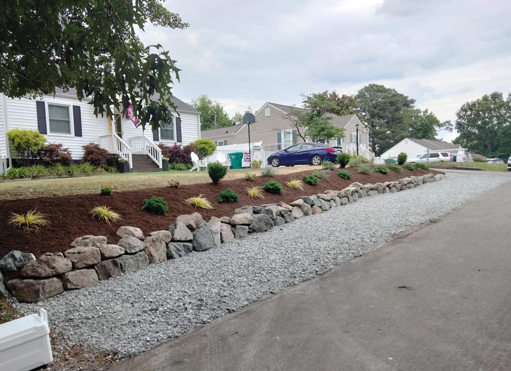 Stone retaining wall with planted shrubs and gravel driveway in front of suburban homes.