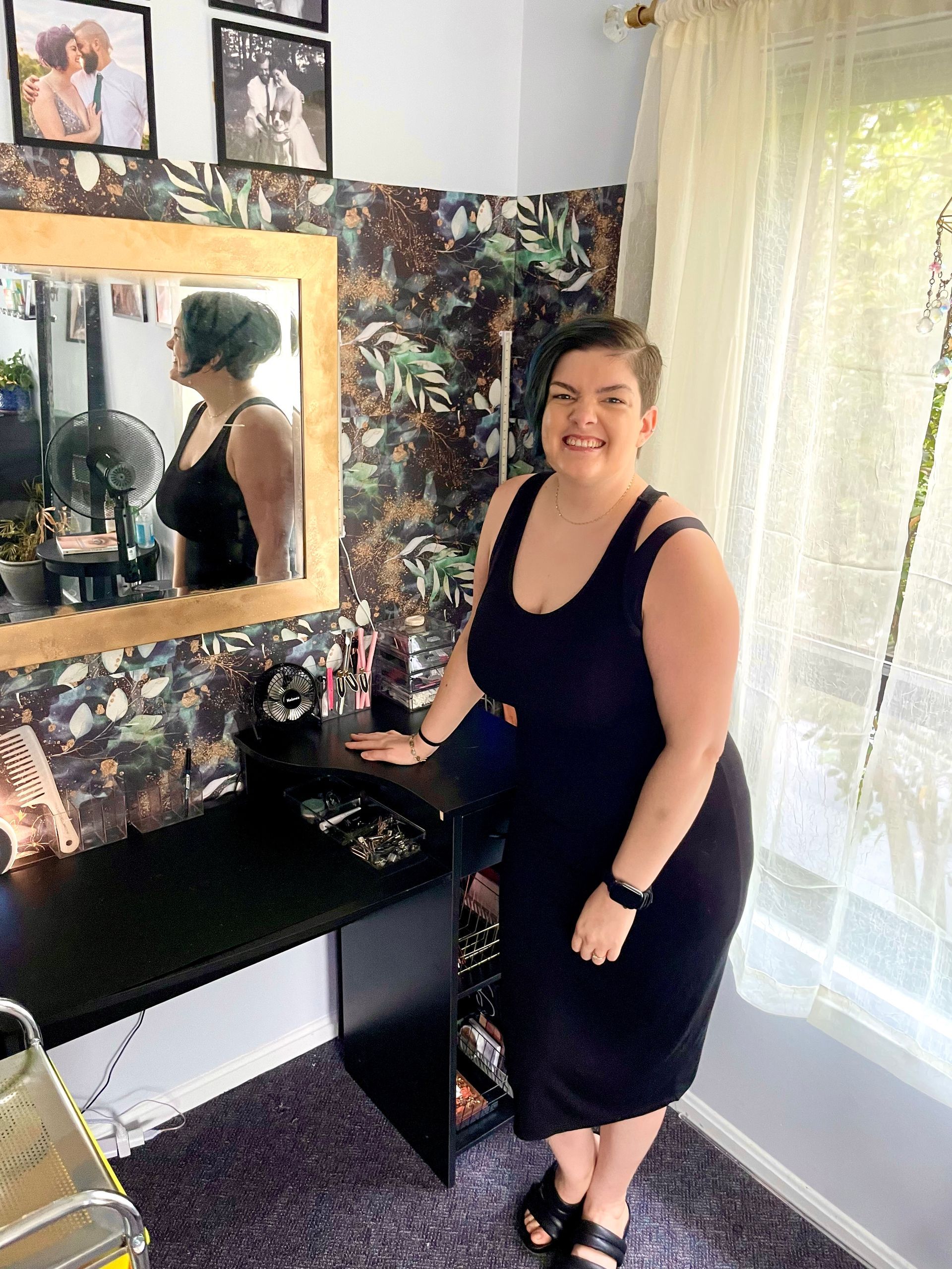 A woman in a black dress is standing in front of a mirror in a salon environment. 