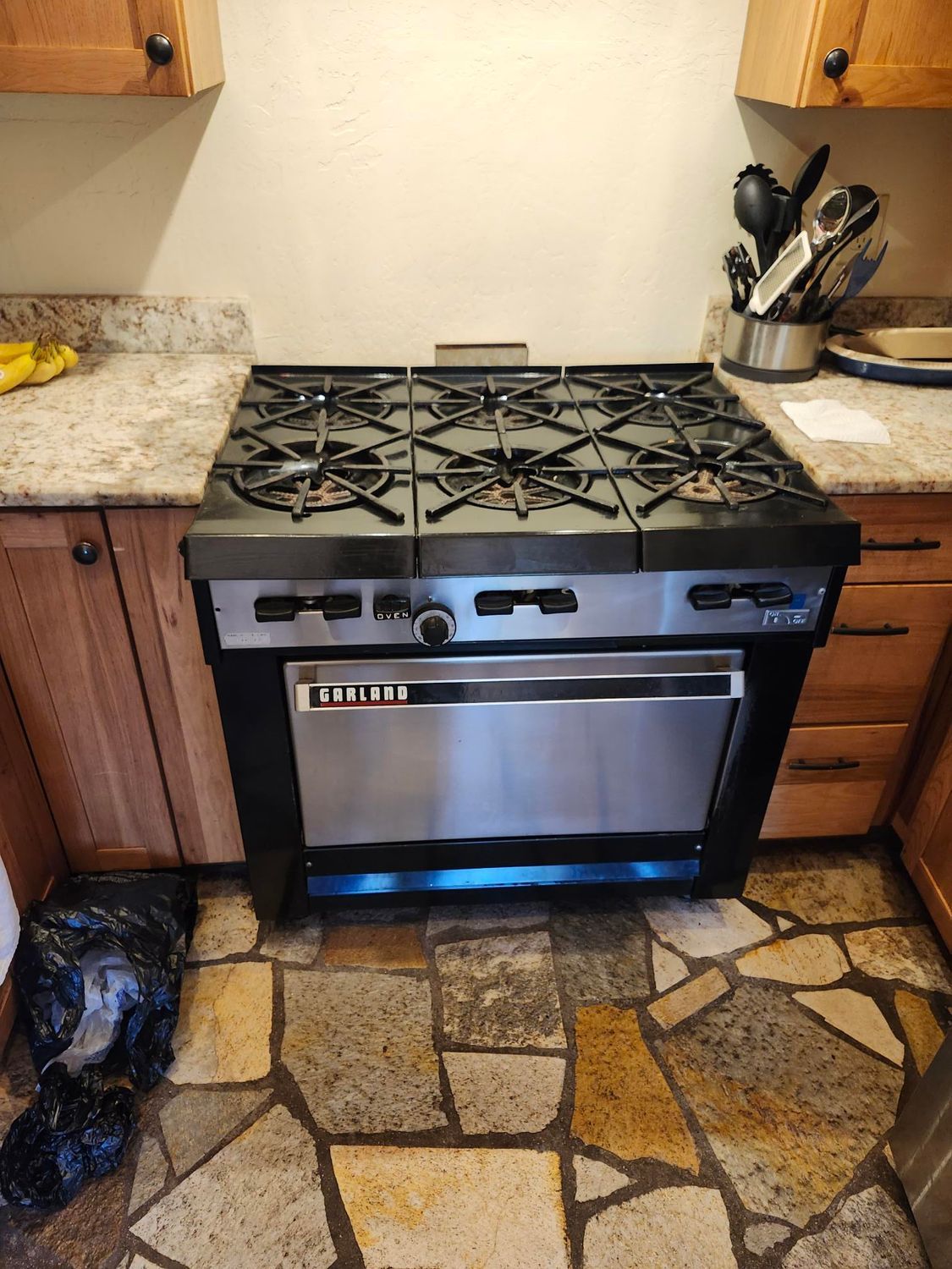 Kitchen stove with six burners and oven, set in wooden cabinets. Stone tile floor.