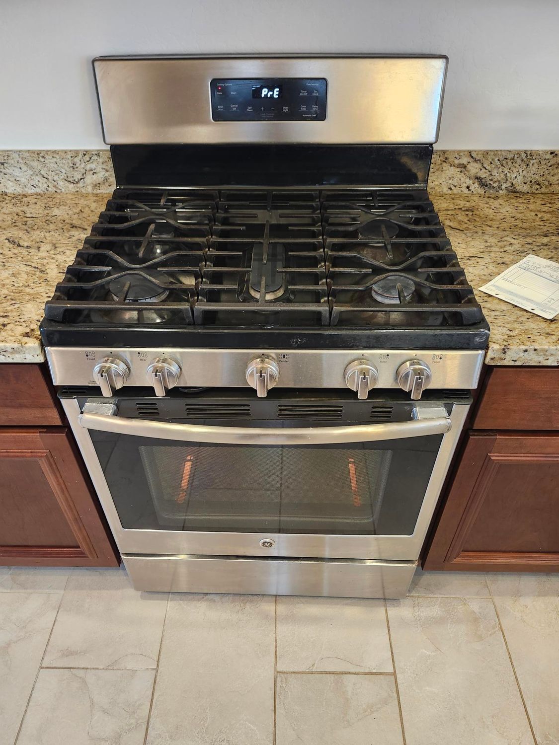 Stainless steel gas range in a kitchen setting with tan countertop and brown cabinets.