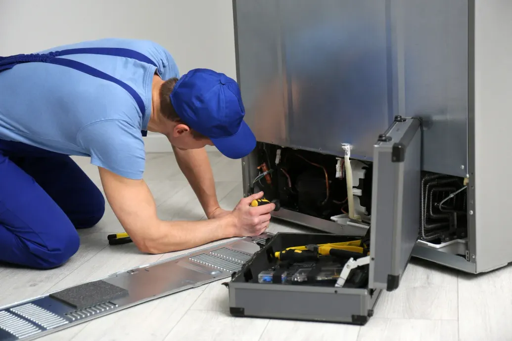 A man is kneeling on the floor fixing a refrigerator.