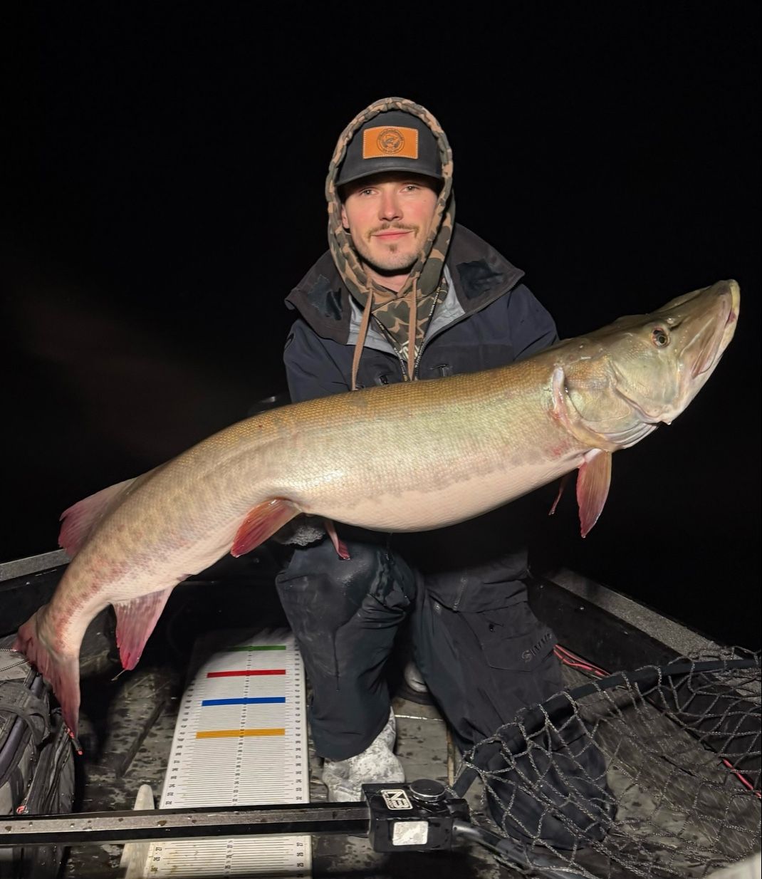 Man holding large fish on a boat at night. He is wearing a winter coat.