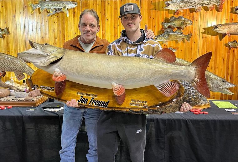 Two people holding a large mounted fish trophy at an event. They stand in front of wood paneling with other mounted fish.
